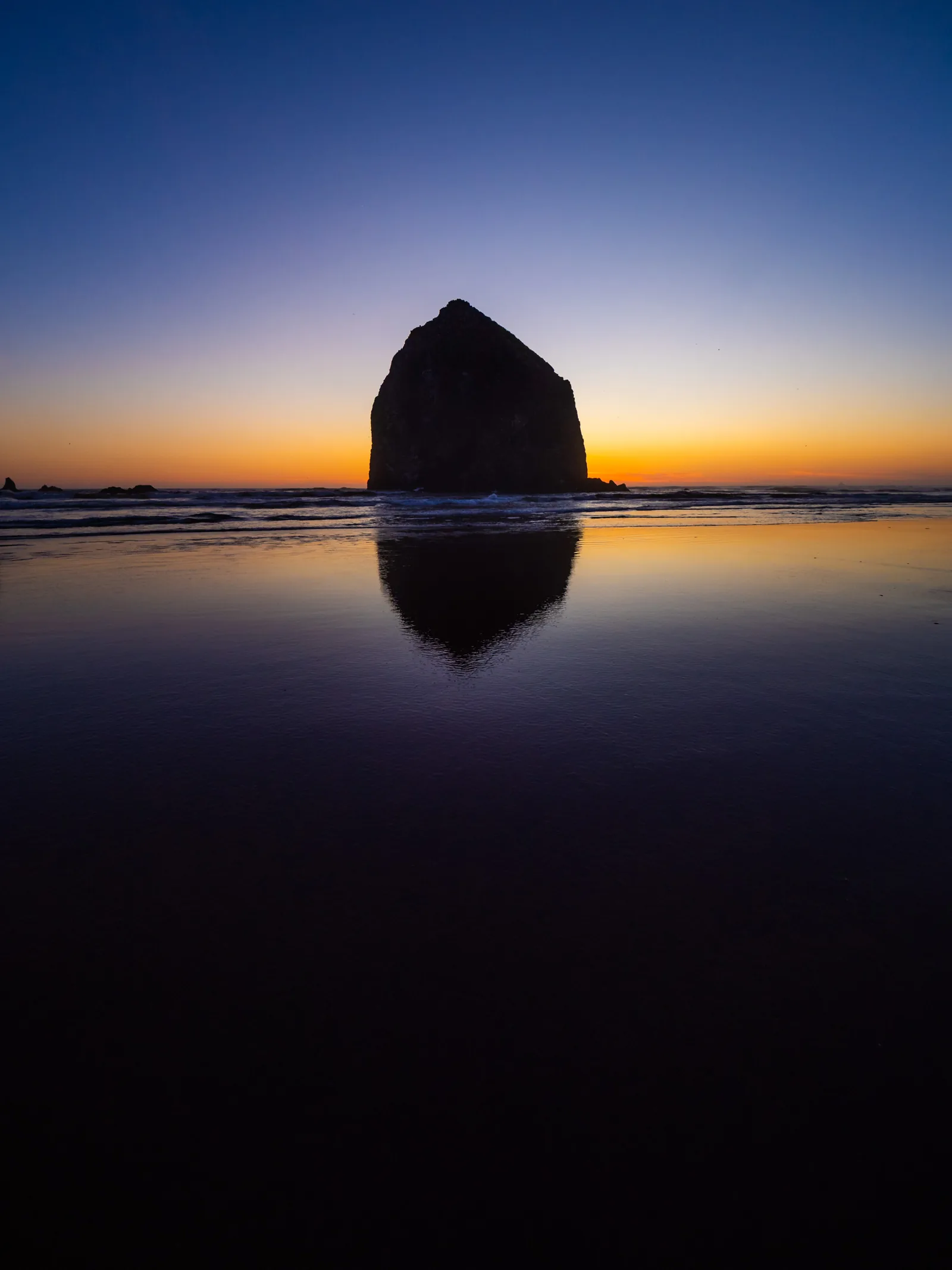 Haystack Rock silhouetted against an orange horizon, reflected in the wet sand.