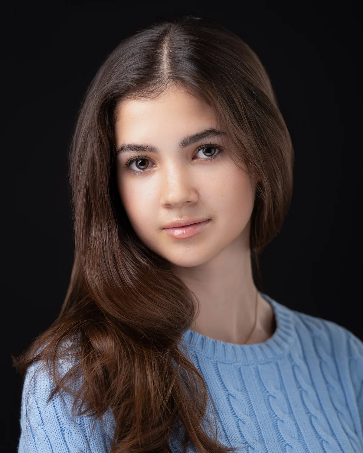 Headshot of a young performer with long brown hair in a light blue cable-knit sweater, against a black background.