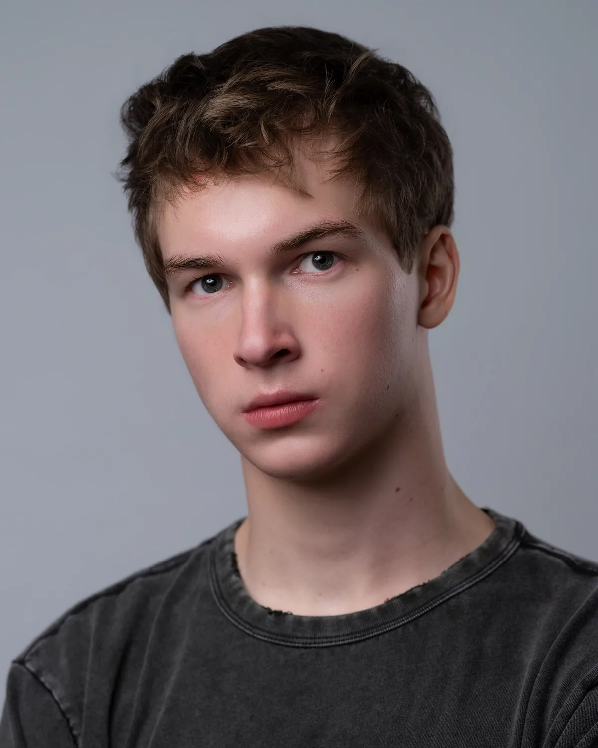 Headshot of a young performer with tousled brown hair in a charcoal T-shirt, against a grey background.