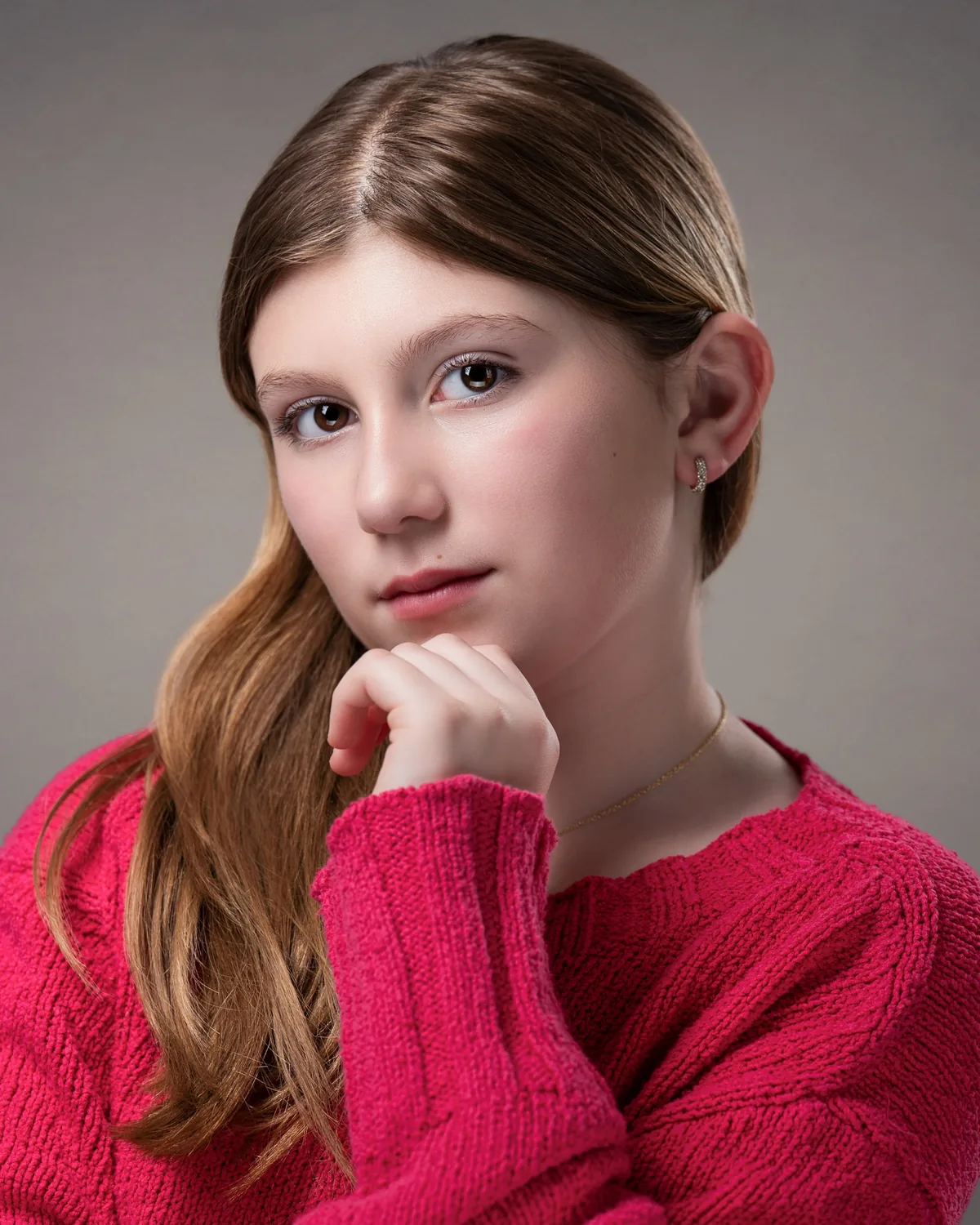Headshot of a young performer with long light-brown hair in a red sweater, chin resting on one hand, against a warm grey background.
