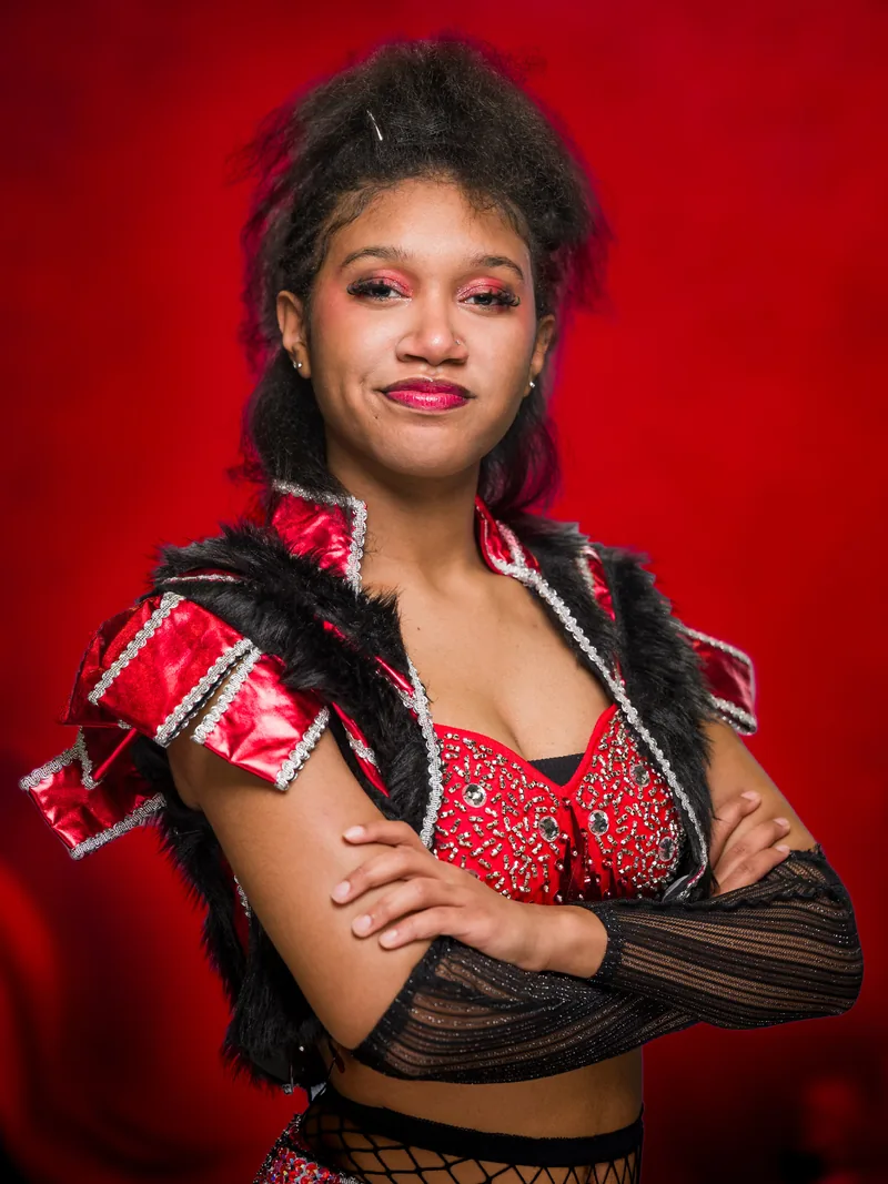 Character portrait of Anna of Cleves from SIX, arms crossed in a red costume against a red background.