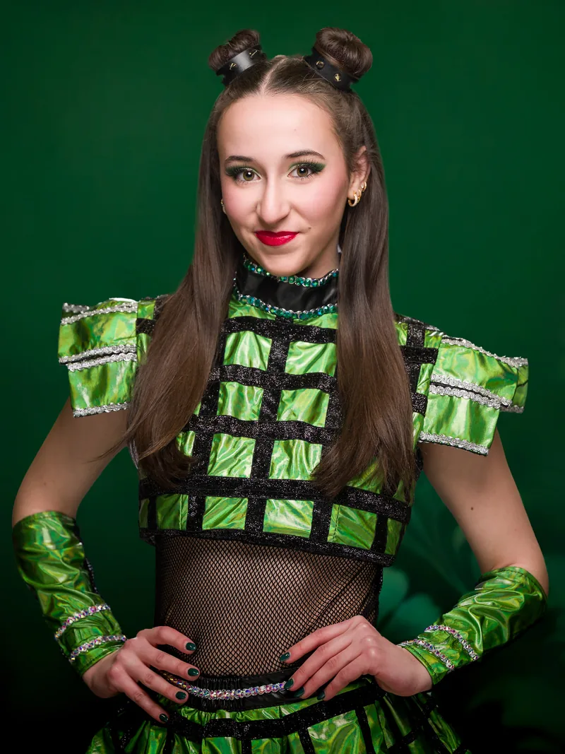 Character portrait of Anne Boleyn from SIX, hands on hips in a green costume against a deep green background.
