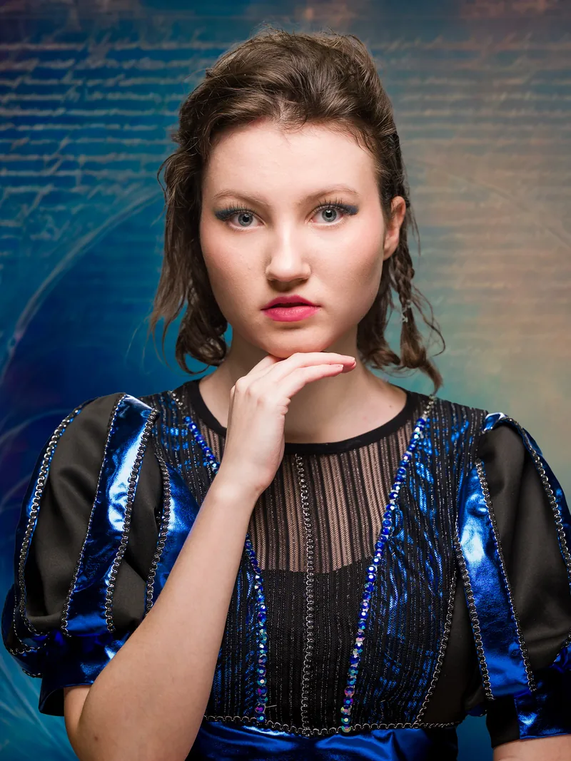 Character portrait of Catherine Parr from SIX, hand at chin in a blue costume against a blue background lined with handwriting.