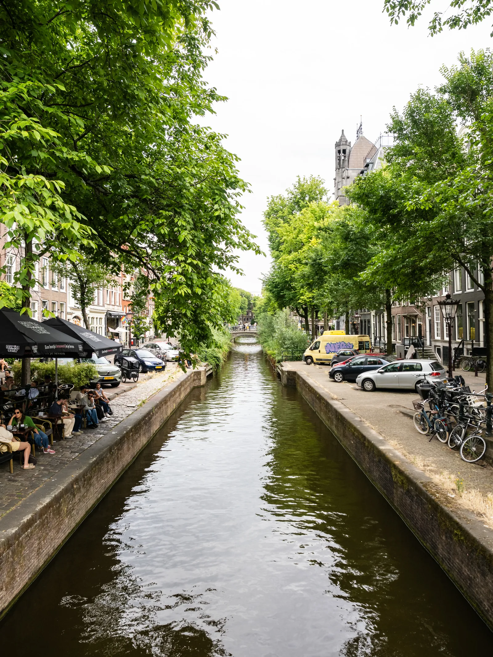 A tree-lined canal with parked cars, bicycles and a café terrace along its banks.