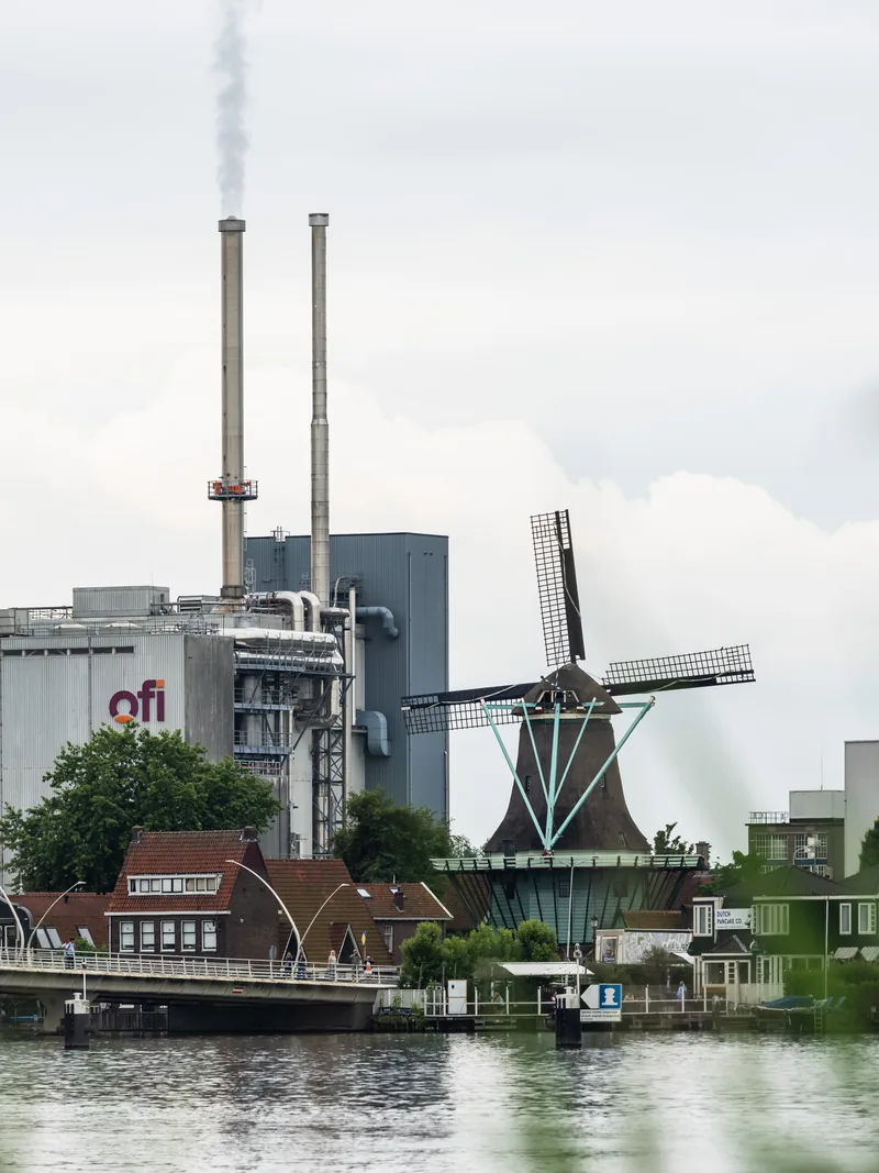 A windmill beside the river in front of an industrial plant with tall smokestacks.
