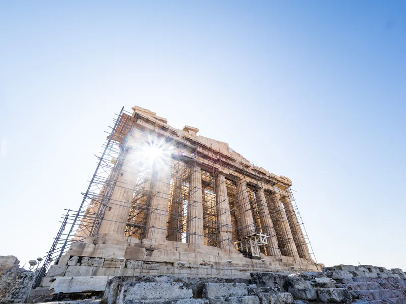 The Parthenon wrapped in restoration scaffolding, with the sun bursting through its columns.