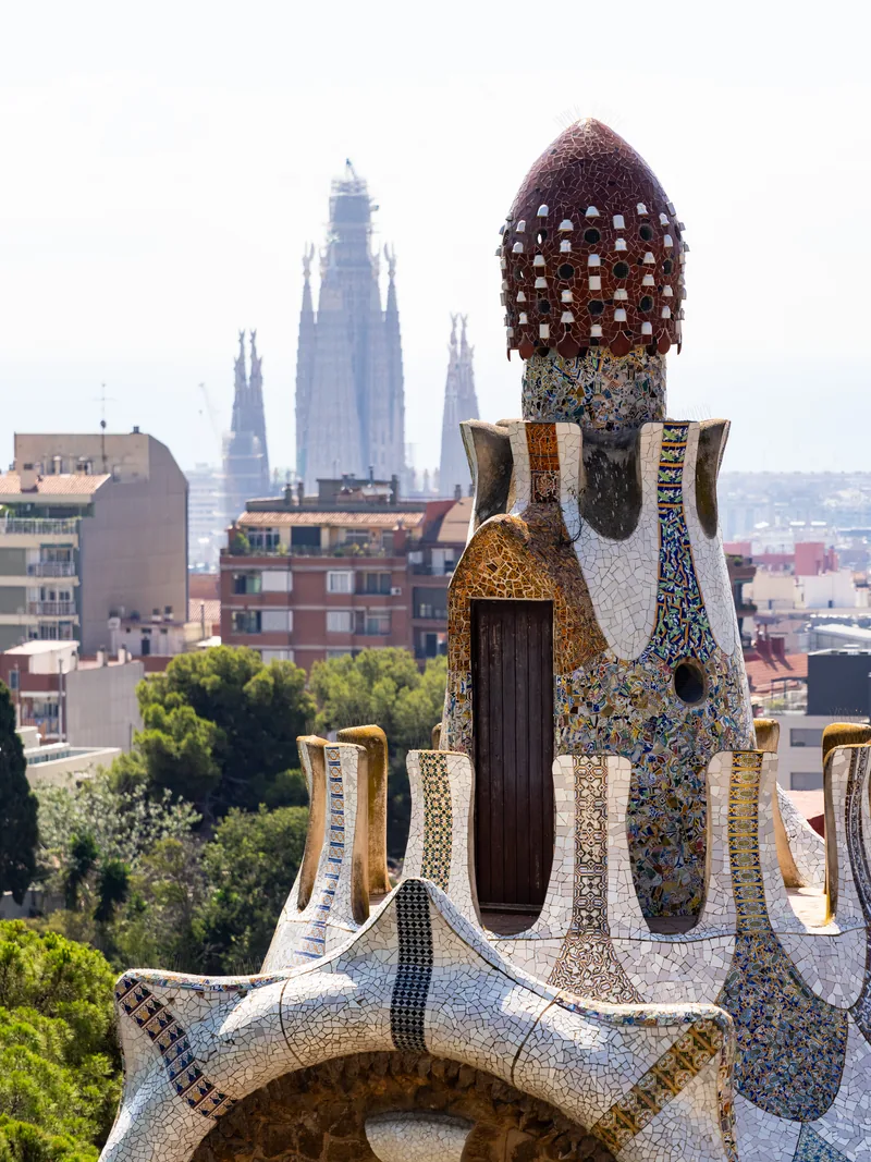 A mosaic-covered Gaudí tower at Park Güell, with Sagrada Família's spires rising in the distance.