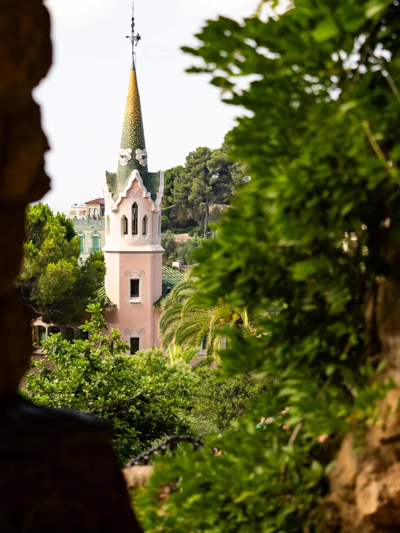 A pink house with a green-tiled spire seen through dark foliage at Park Güell.