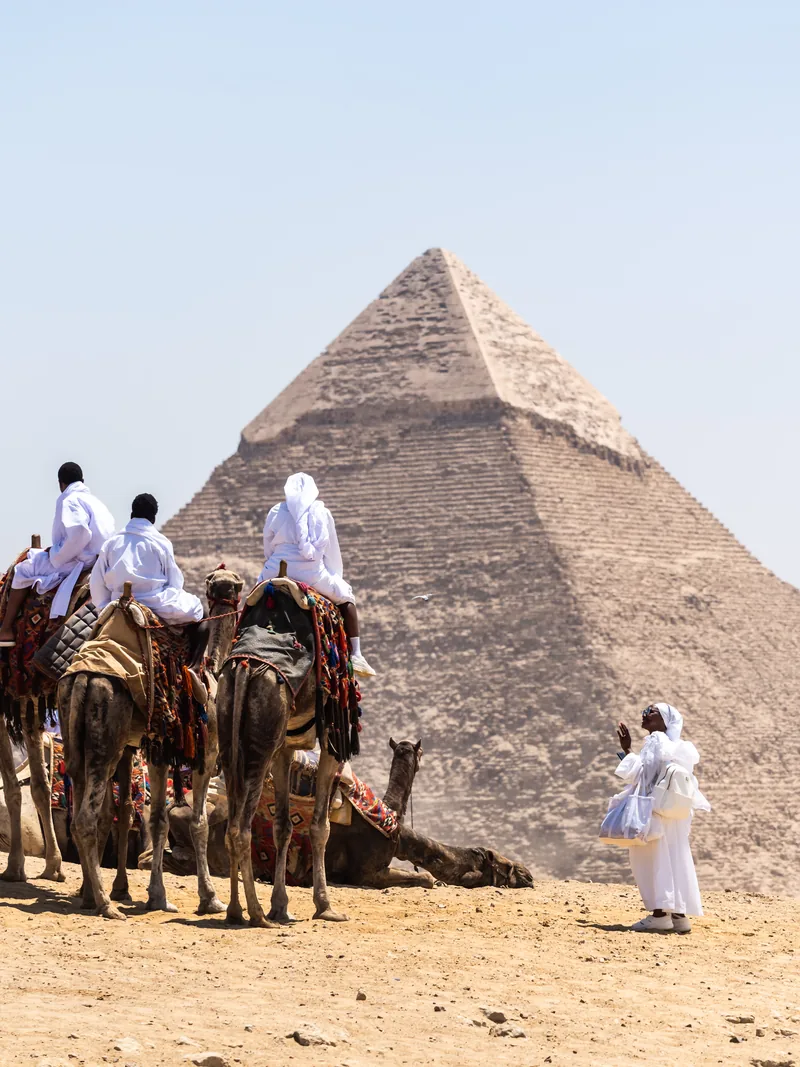 Camel riders in white robes resting in front of a pyramid.