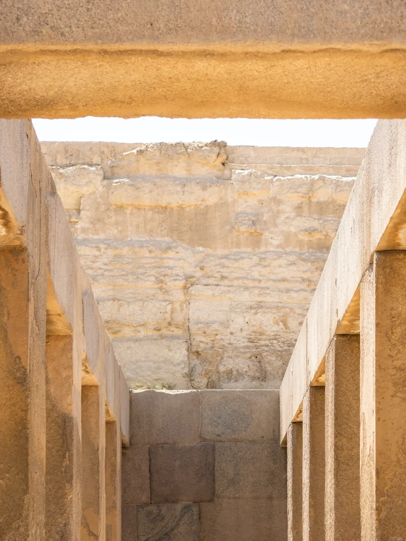 A stone passageway with square columns leading toward a sunlit wall of rock.