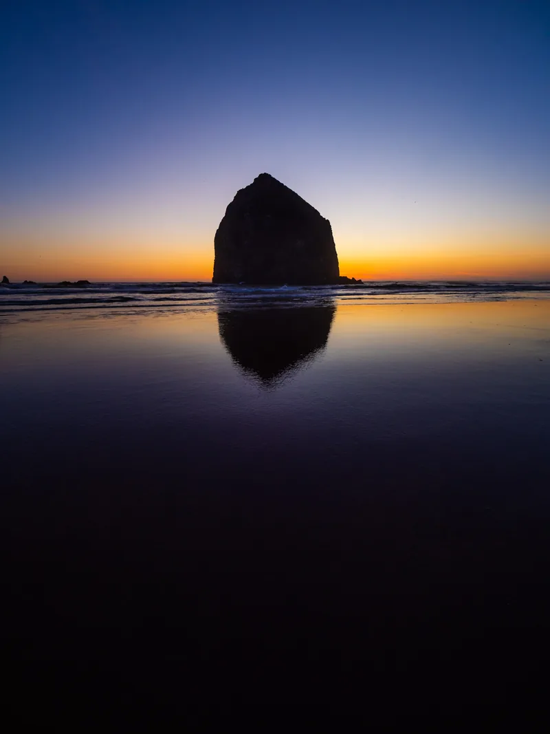 Haystack Rock silhouetted against an orange horizon, reflected in the wet sand.