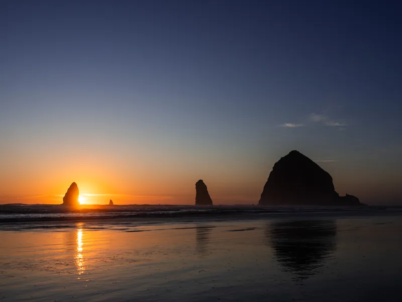 The sun setting between sea stacks at Cannon Beach, reflected in the sand.