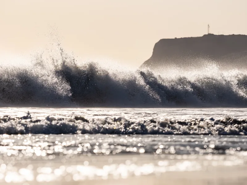 A wave breaking in backlit spray with Point Loma behind.