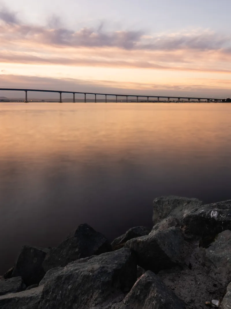 The Coronado Bridge on the horizon above still water and dark rocks at dawn.