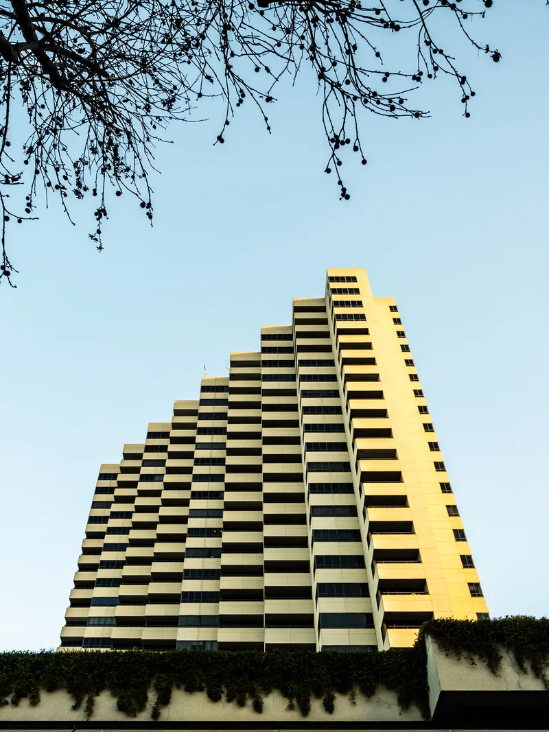 A stepped yellow hotel tower against a pale blue sky, framed by branches.