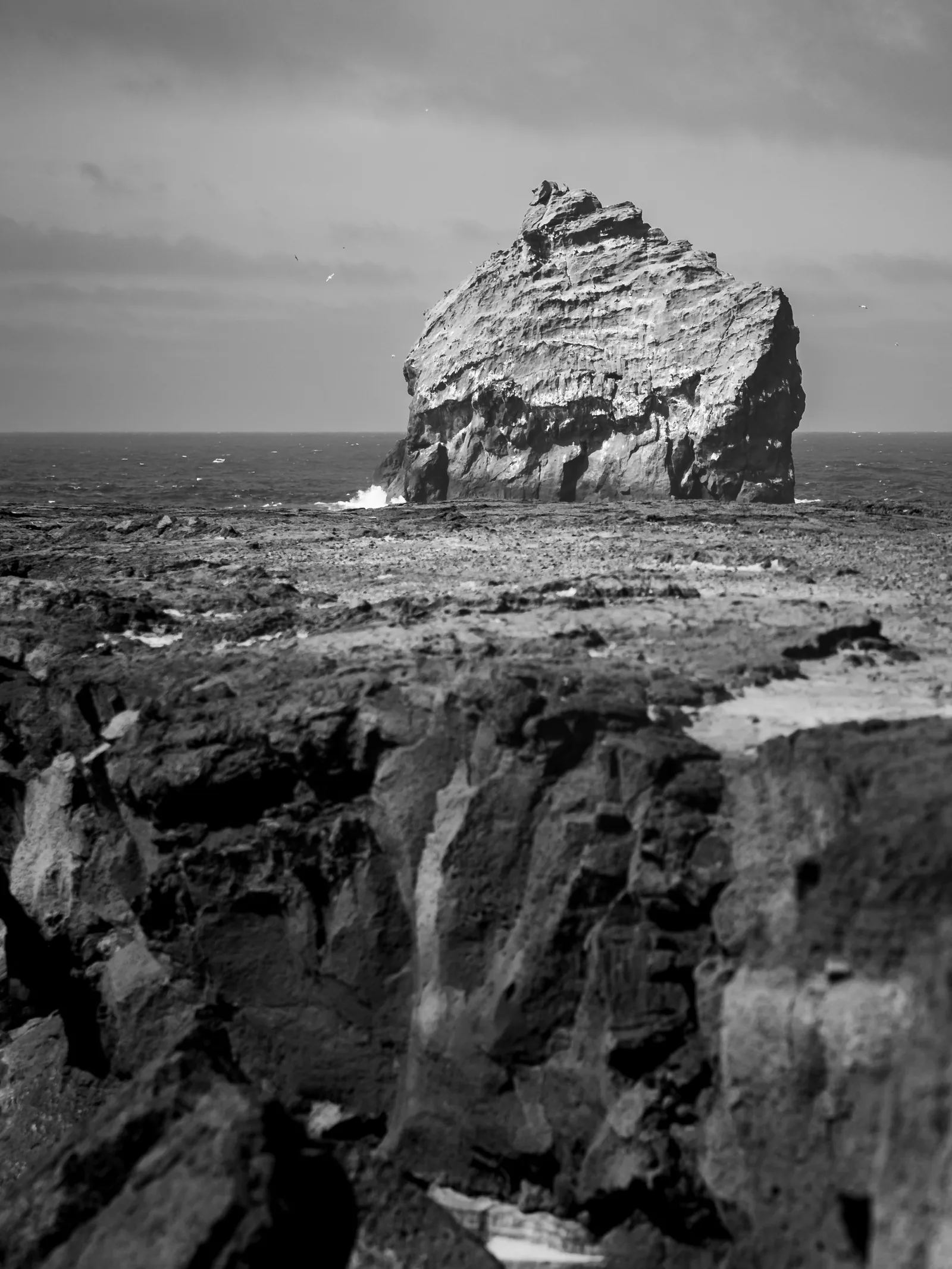 A sea stack off the Icelandic coast beyond dark lava cliffs, in black and white.