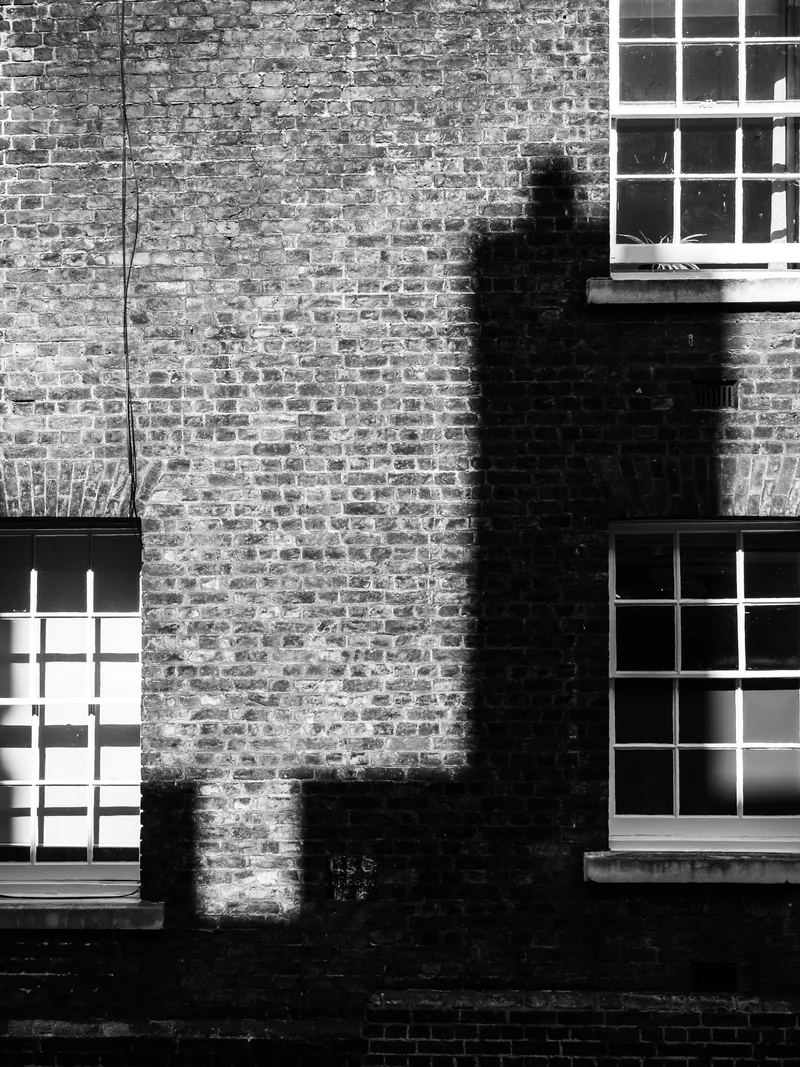 A stepped shadow across a London brick wall between white window frames, in black and white.