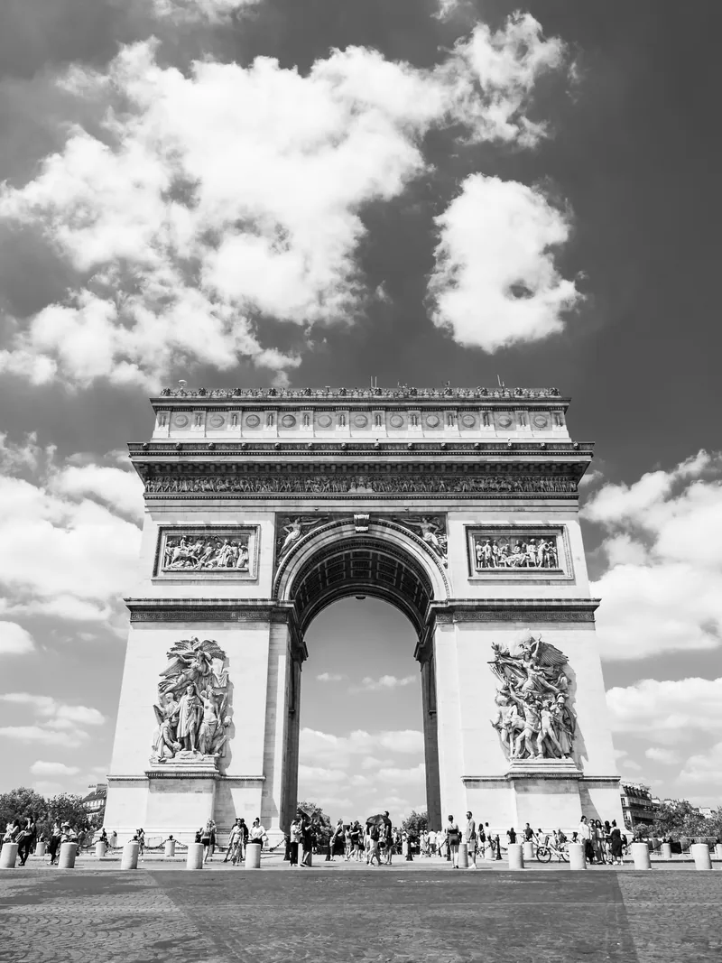 The Arc de Triomphe under bright clouds, with crowds at its base, in black and white.
