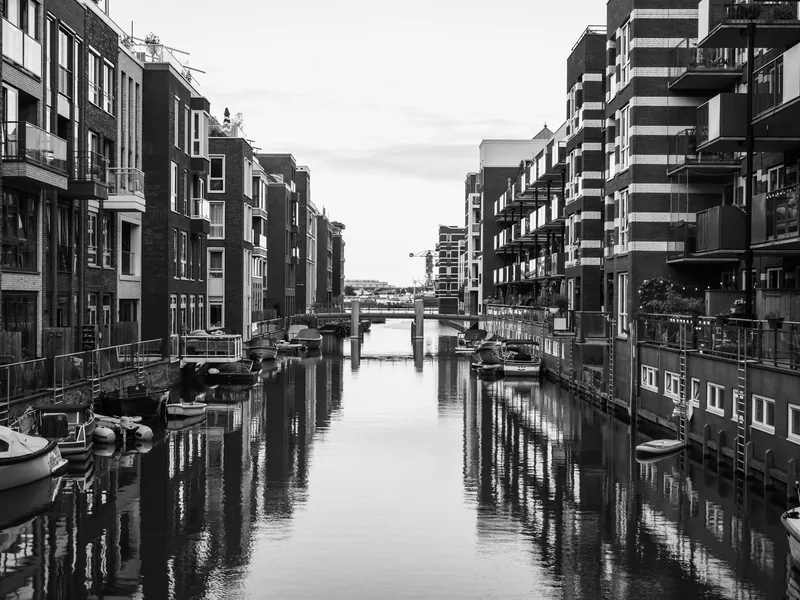 A canal between modern Amsterdam apartment blocks reflected in still water, in black and white.
