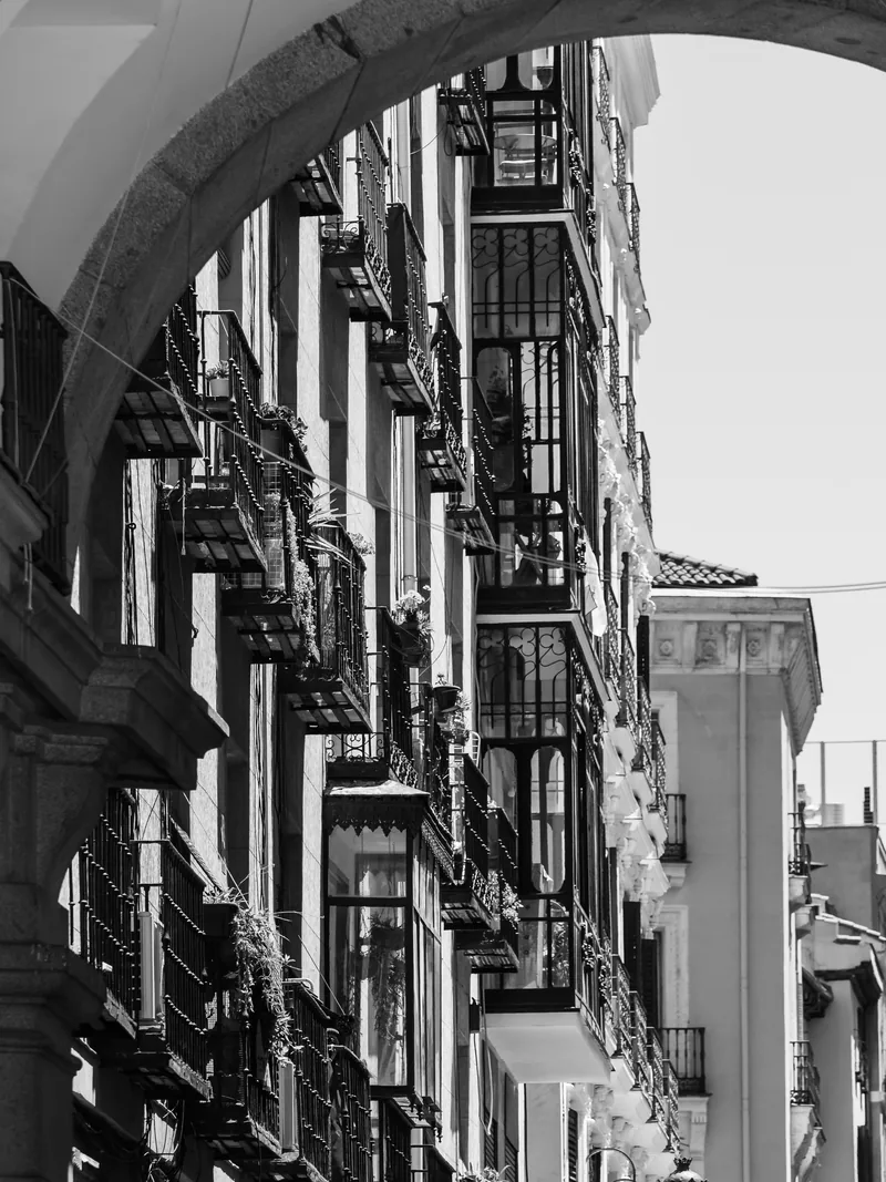 Balconies climbing a narrow Madrid street seen through a stone arch, in black and white.