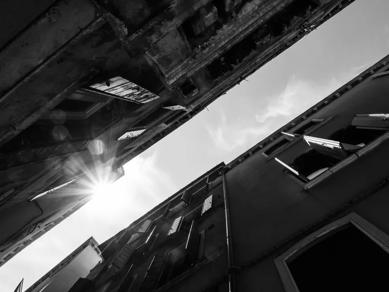 The sun bursting between two leaning Venetian façades, in black and white.