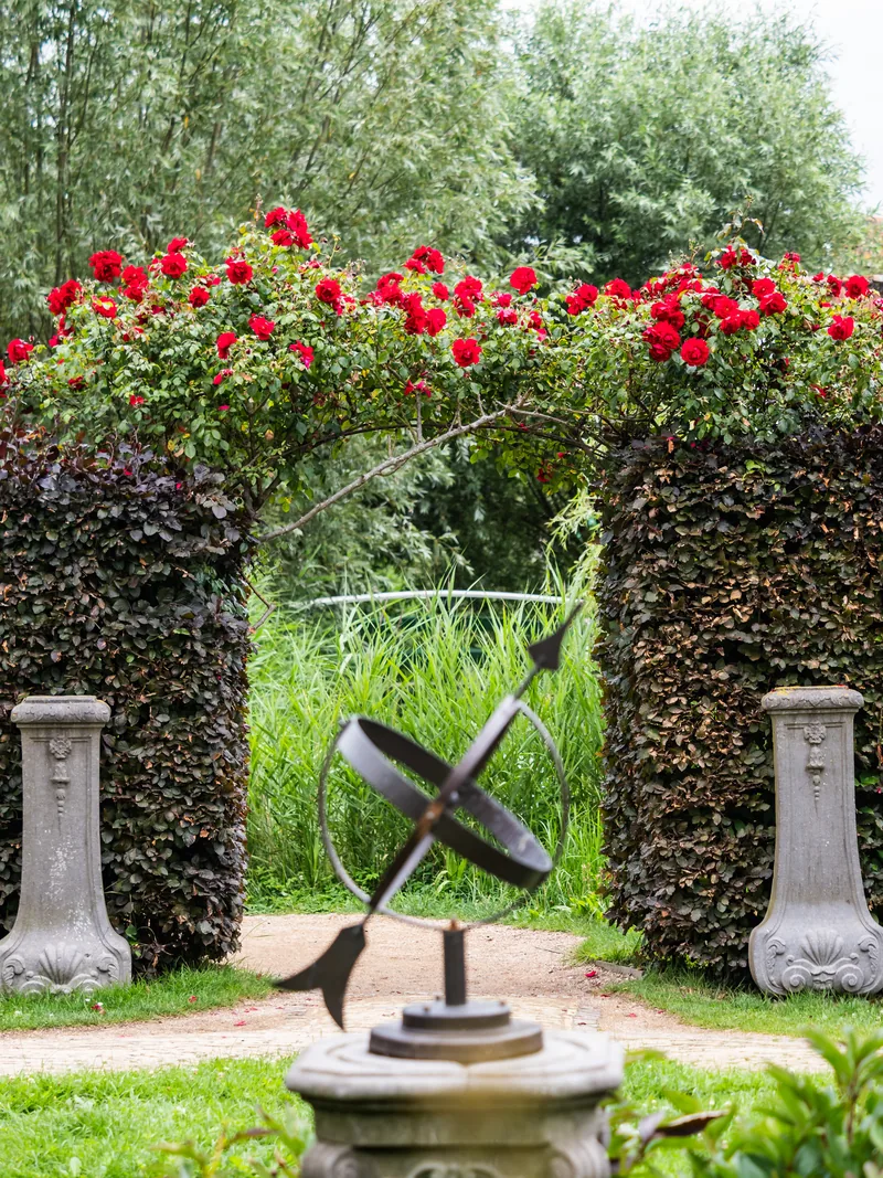 An armillary sphere sundial in front of an archway of hedge and red roses.