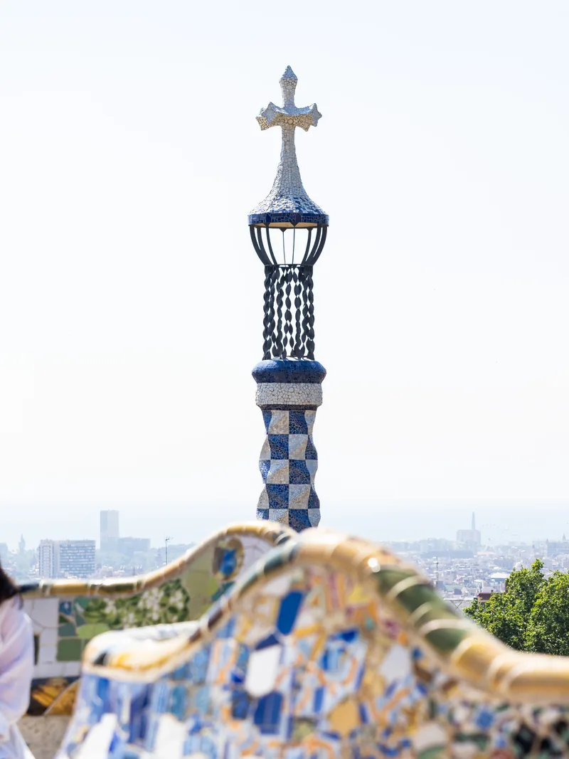 A mosaic spire topped with a cross above Park Güell's wavy tiled bench, the city in the haze below.