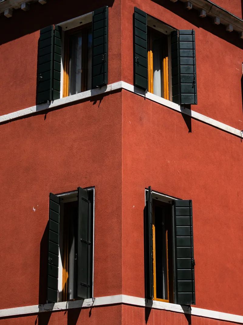 A red Venetian wall with dark green shutters on the corner of a building.