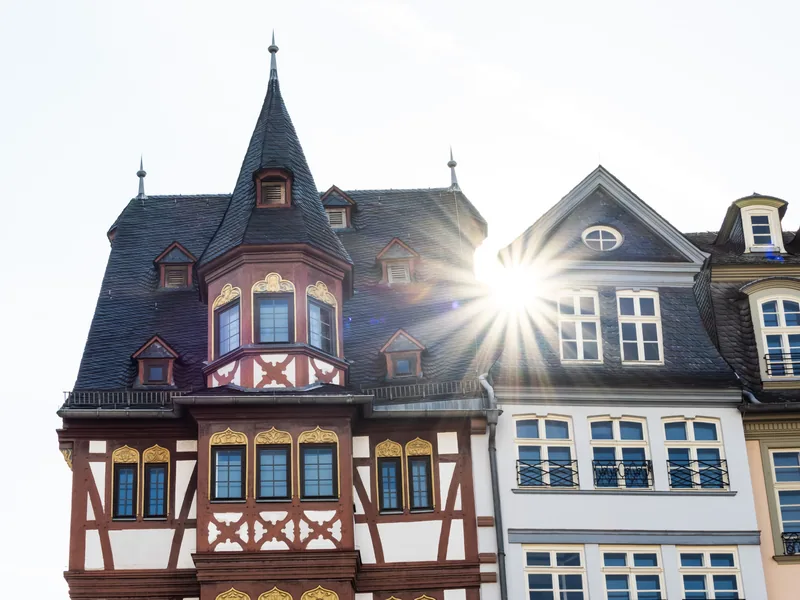 A half-timbered house with a slate turret beside white townhouses, the sun flaring behind a gable.