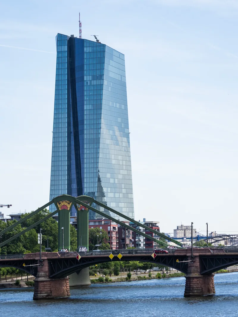The glass tower of the European Central Bank rising behind a green iron bridge over the Main.