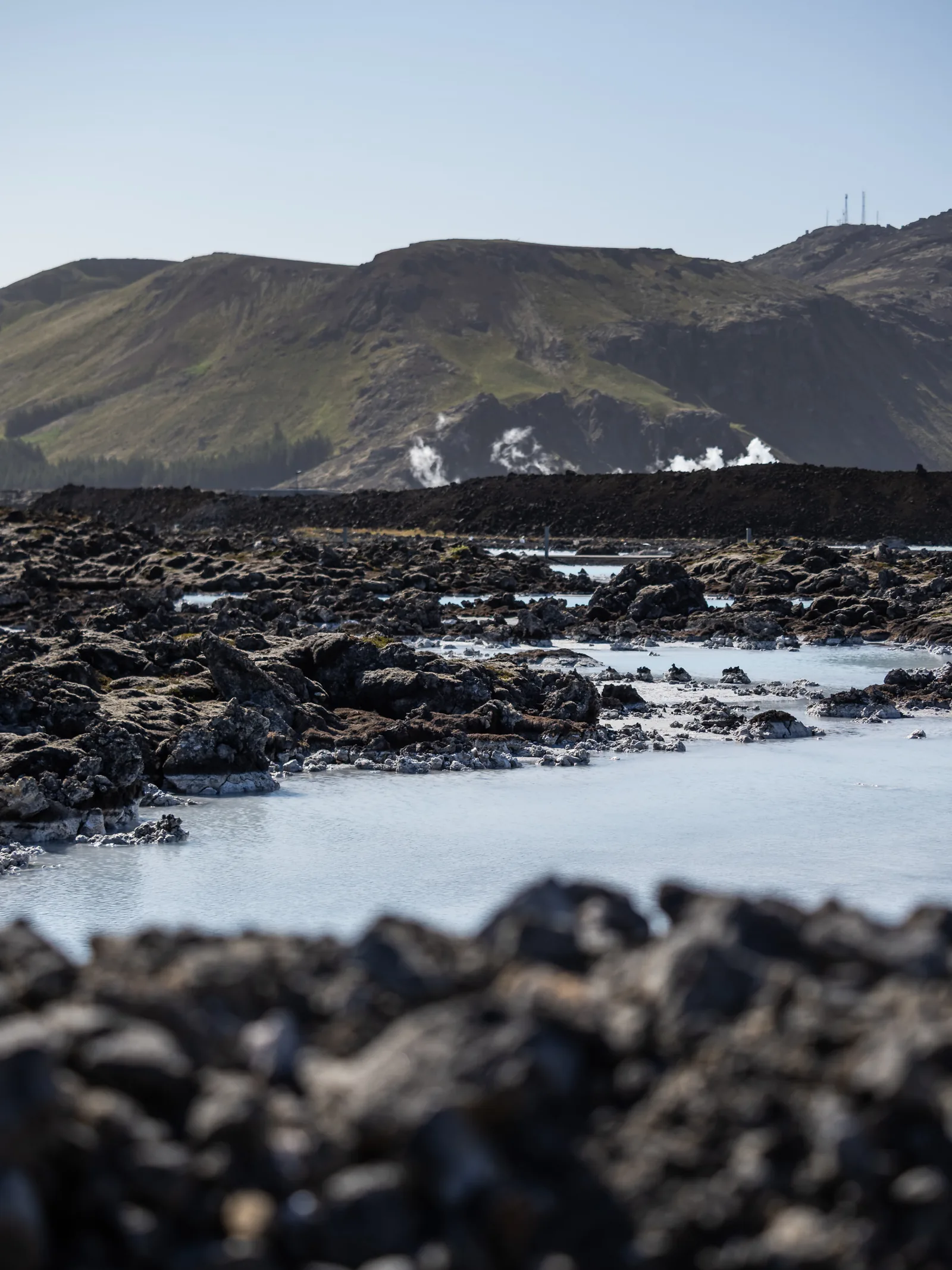 Milky blue geothermal water between black lava rocks, with steam rising at the foot of a green hillside.