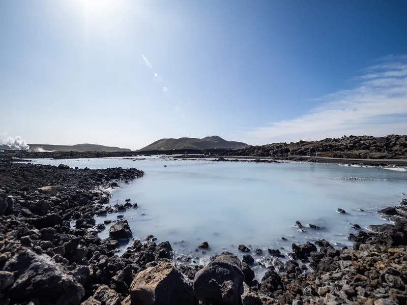 A pale blue geothermal lagoon ringed by black lava rock under a bright, hazy sun.