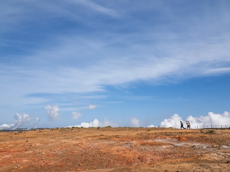 Two walkers crossing a rust-orange geothermal plain as plumes of steam rise along the horizon.