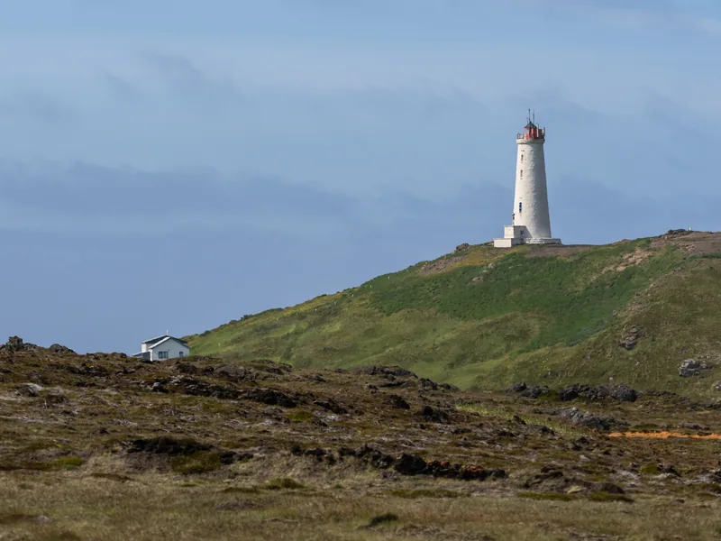 A white lighthouse with a red top on a green hill above dark, rocky ground.