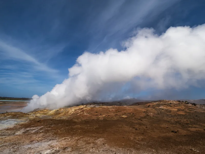 A billowing cloud of geothermal steam rising from orange earth against a streaked blue sky.