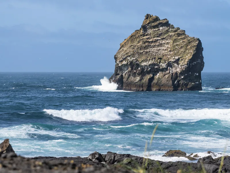 A rugged sea stack off the coast, with waves breaking white around it.