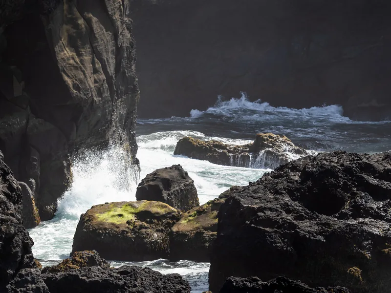 Surf crashing between black volcanic rocks at the base of a sea cliff.