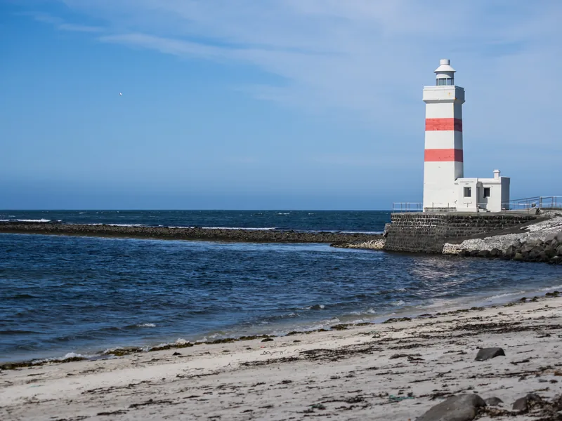 A white lighthouse with red bands at the end of a stone breakwater beside a sandy beach.