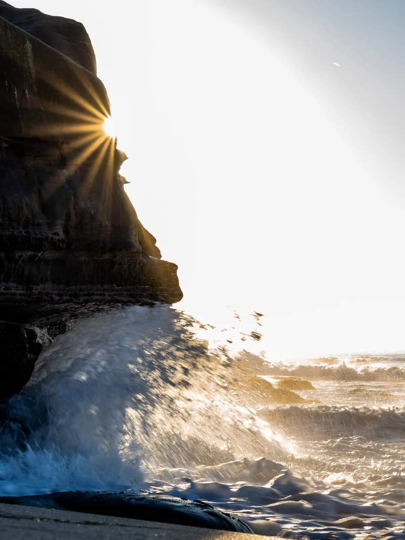 Sunlight flaring past a dark rock as surf crashes below.