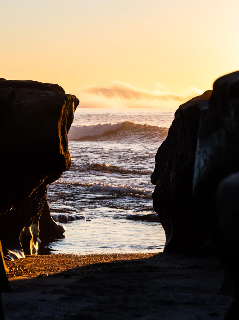 Waves seen between two dark rocks as the sky turns orange.