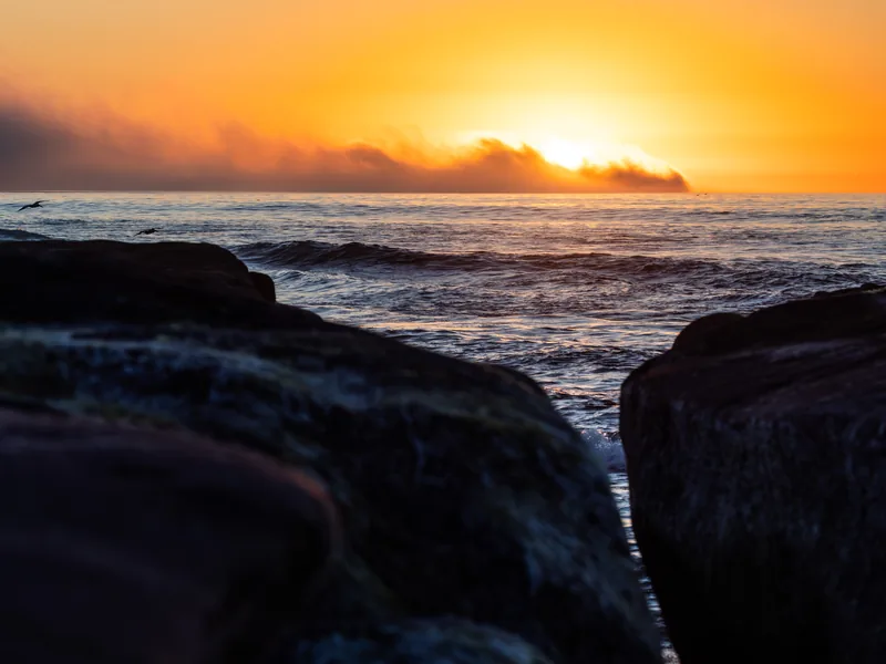 The sun setting behind a bank of clouds above dark rocks and surf.