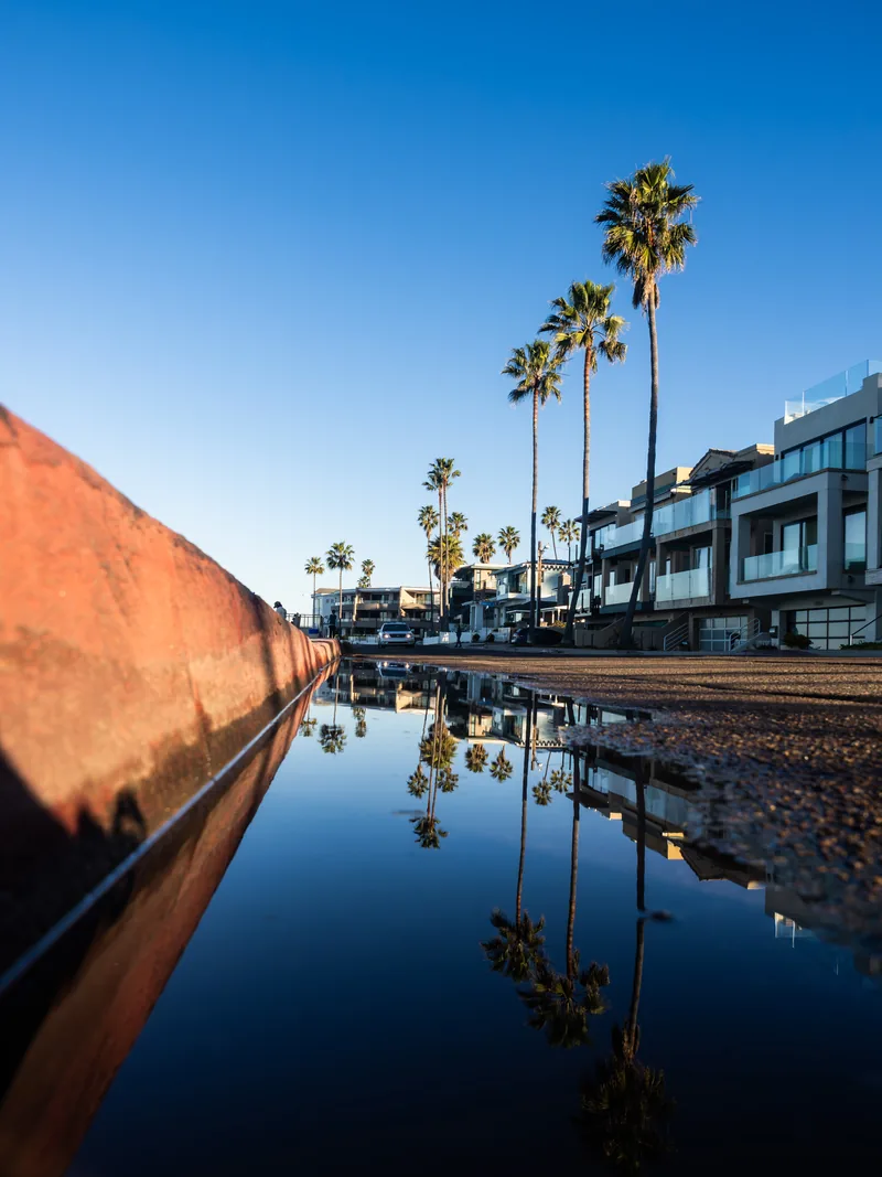 Palm trees and beachfront buildings reflected in a shallow pool beside a red wall.
