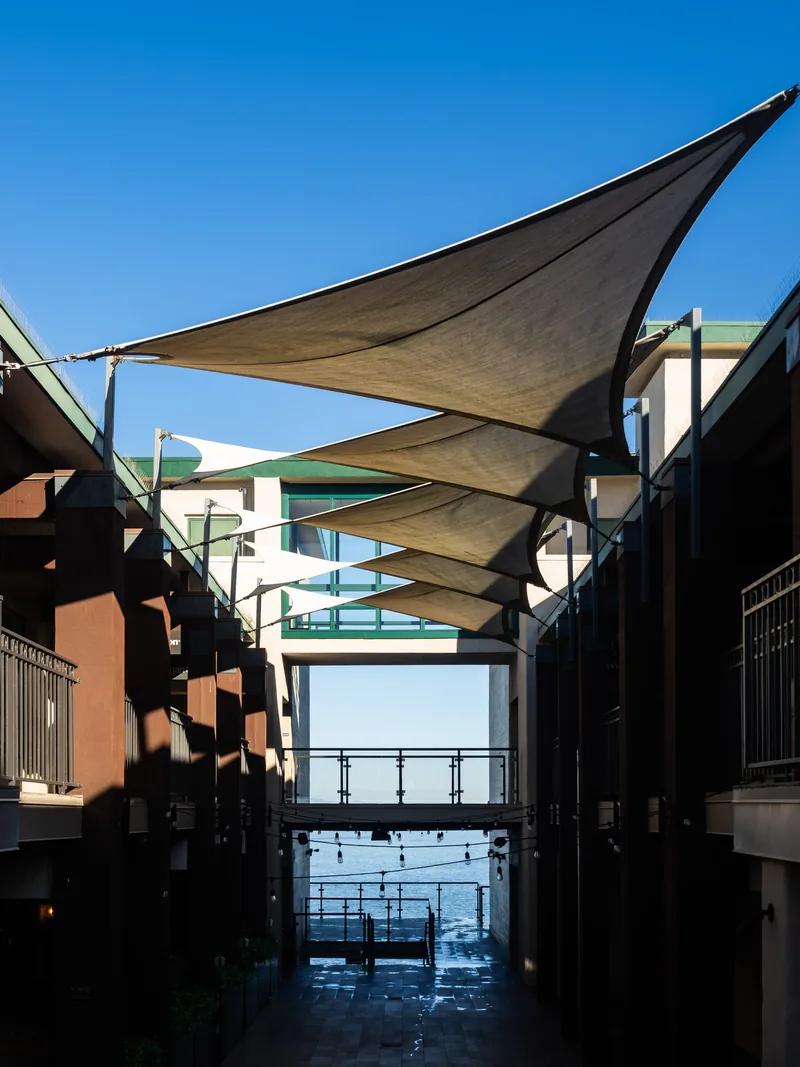 Shade sails over a walkway between buildings, with the ocean at the end.
