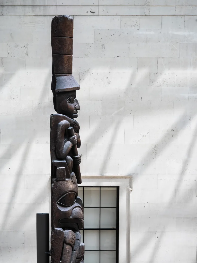 A carved wooden totem pole standing against a pale stone wall streaked with shadows.