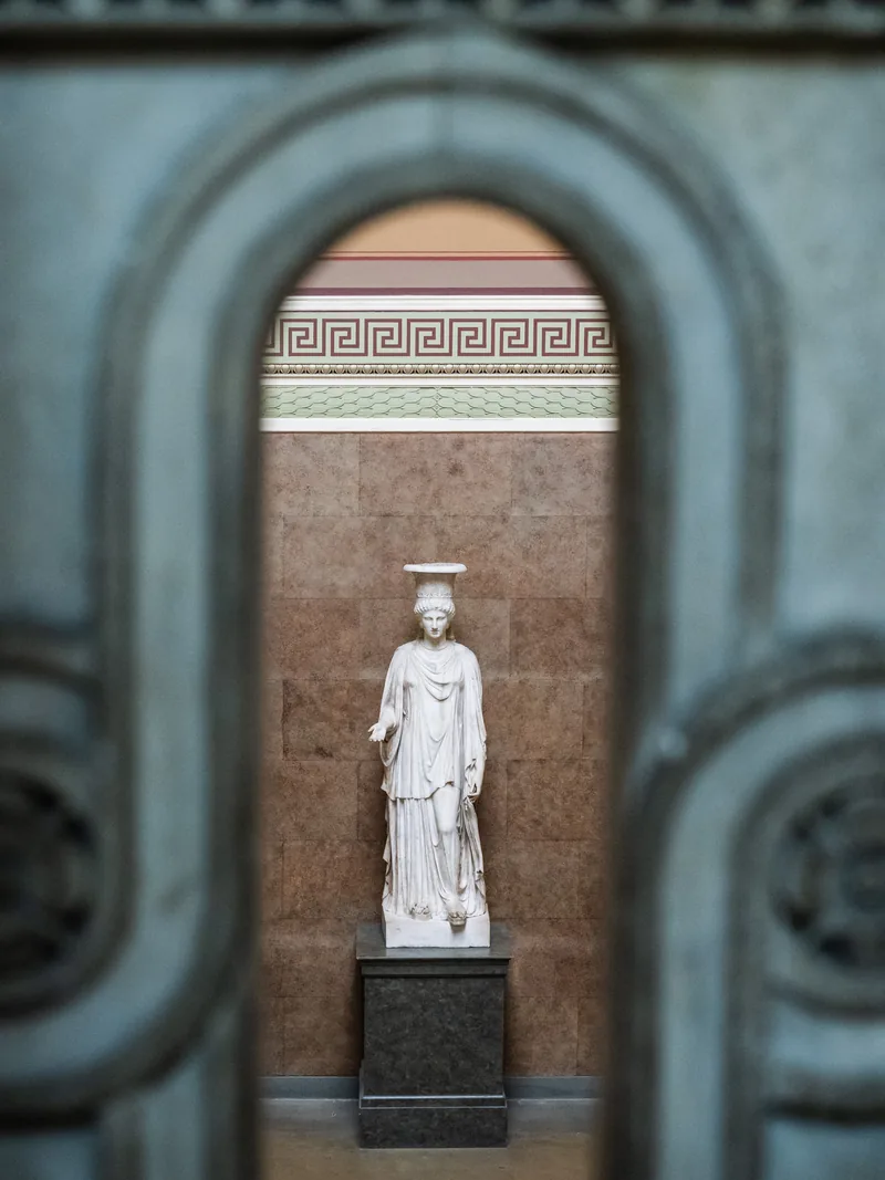 A white classical statue of a woman framed through a softly blurred iron doorway.