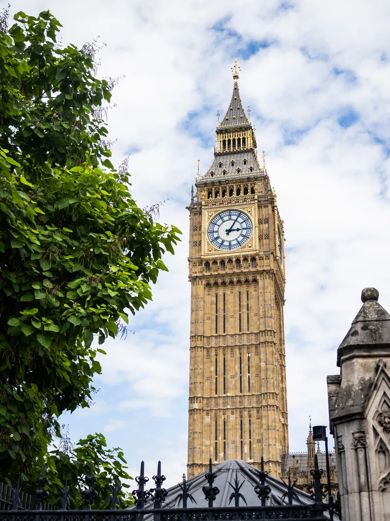 The Elizabeth Tower, home of Big Ben, rising behind trees and iron railings under a cloudy sky.