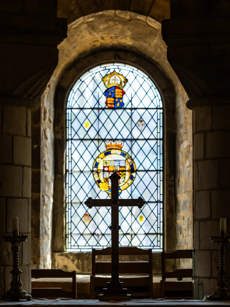 A stained-glass window with royal coats of arms above a simple altar cross in a stone chapel.