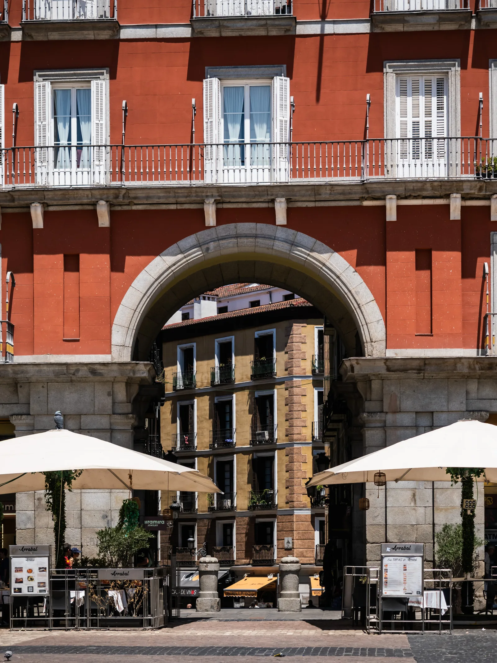A red building with wrought-iron balconies above an arched entrance and café umbrellas on Plaza Mayor.