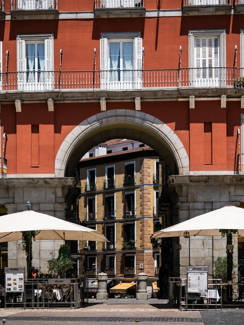 A red building with wrought-iron balconies above an arched entrance and café umbrellas on Plaza Mayor.