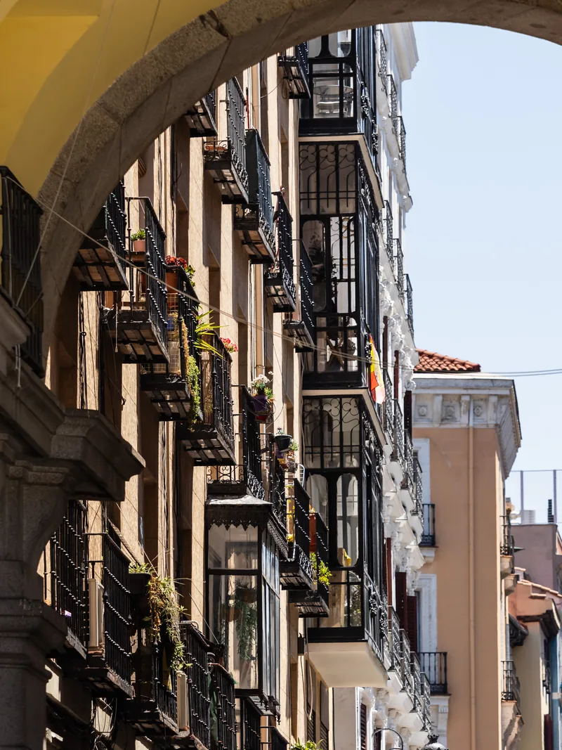 Balconies and hanging plants along a narrow street, framed by a stone archway.